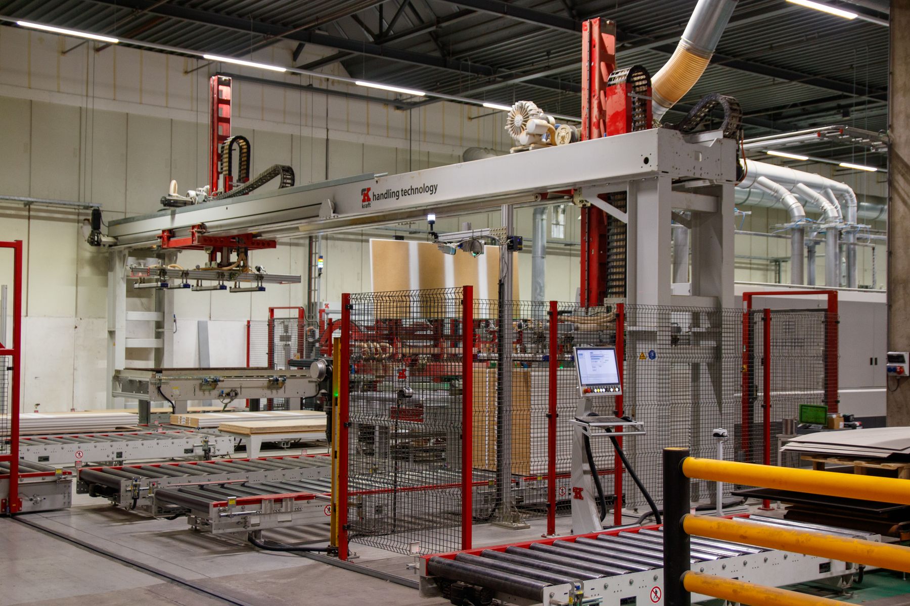 Overhead handling gantry, roller conveyors and stacked flat panels behind mesh fencing.